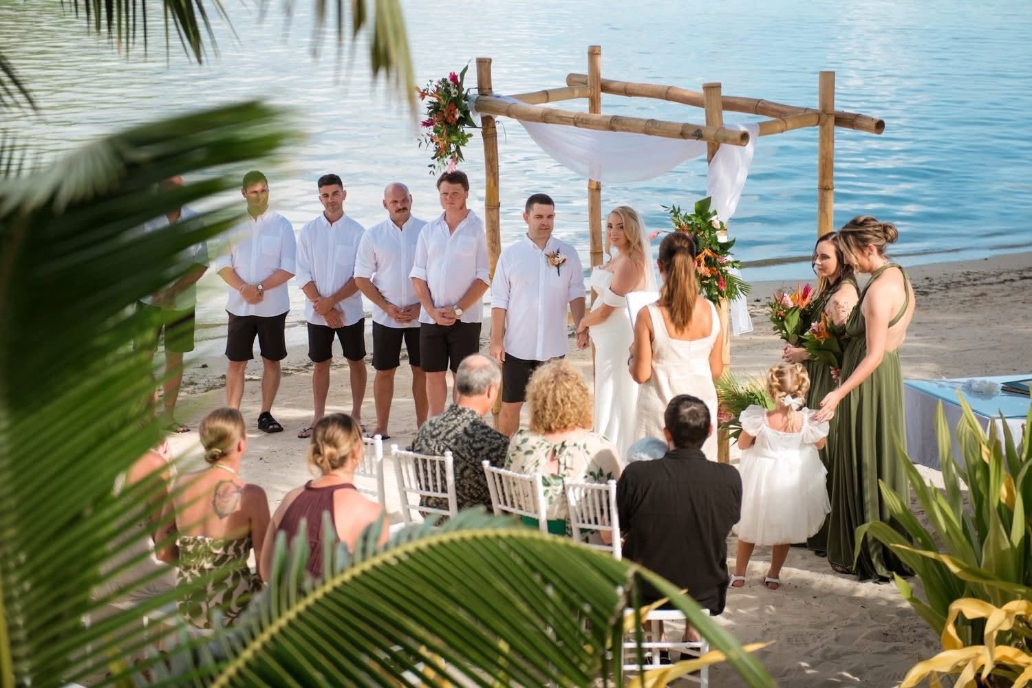 Jane leading a beach ceremony beneath a bamboo arch, with bridesmaids, groomsmen and seated guests