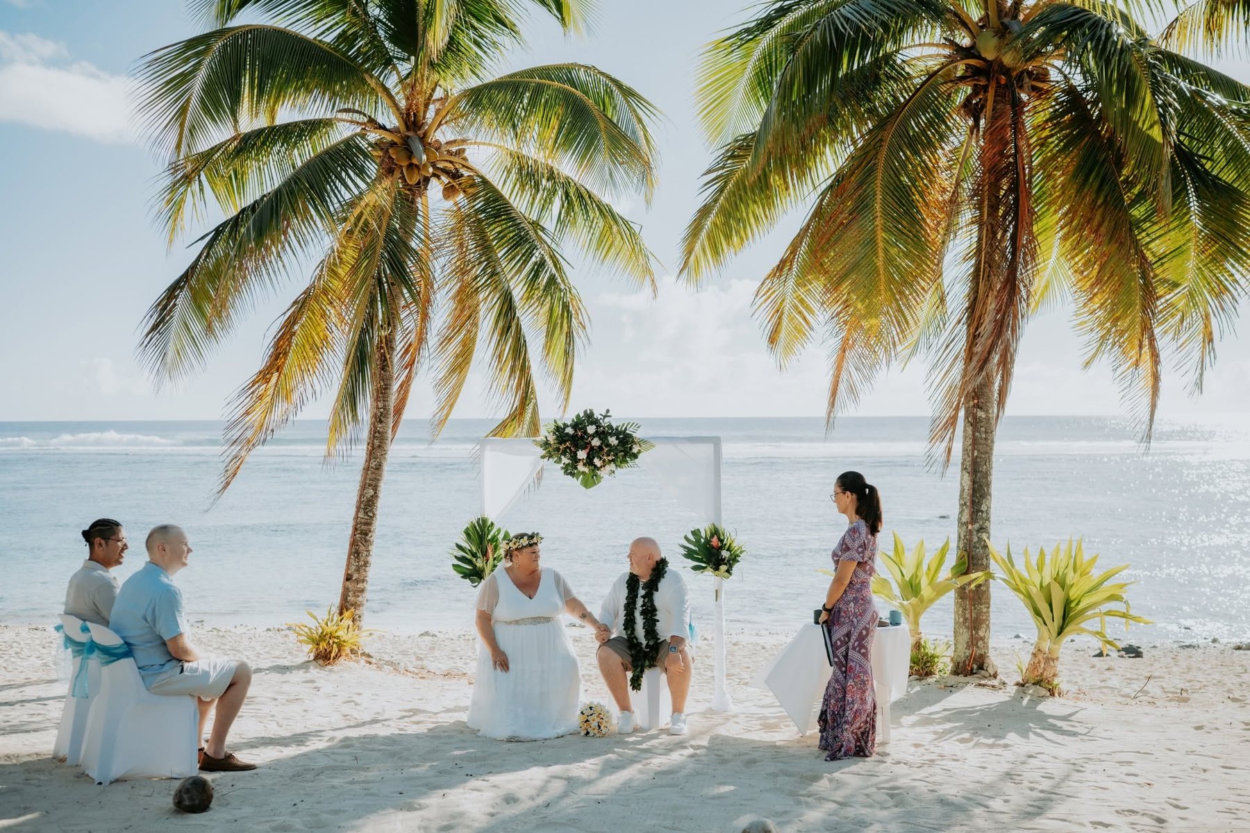 A couple seated beneath coconut palms on a white-sand beach as Jane leads their ceremony