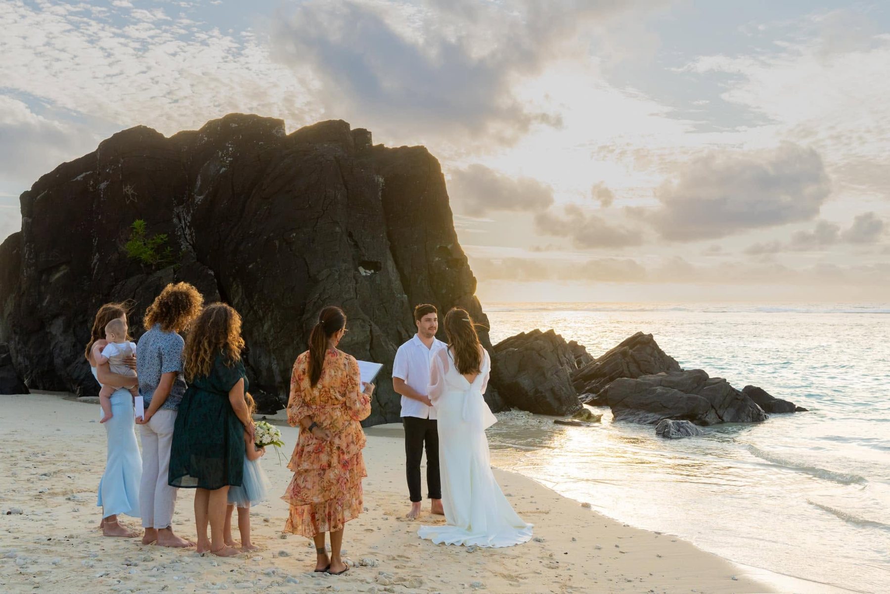 Jane leading a sunset ceremony on the sand beside a tall black rock, family gathered close